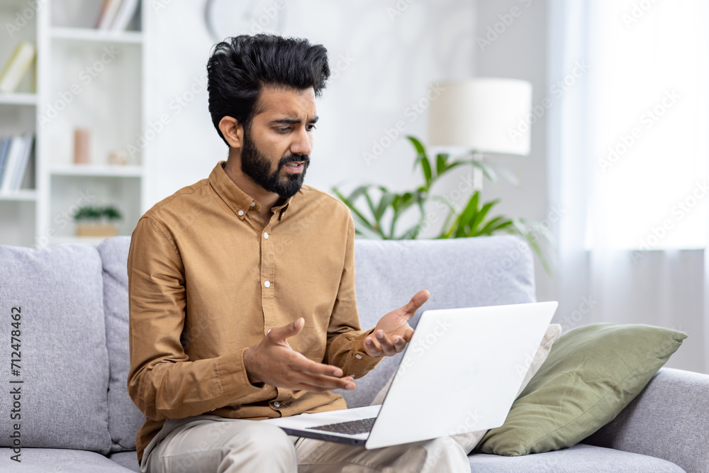 Focused Indian man on sofa with laptop in a cozy living room, capturing modern home life and remote work. Focused Indian man on sofa with laptop in a cozy living room, capturing modern home life and remote work.