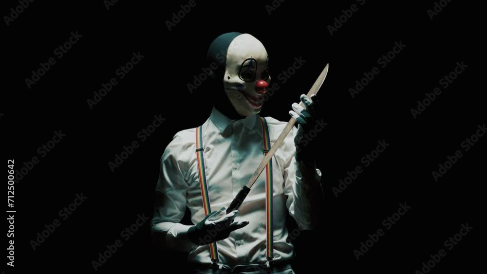 Killer clown looking at a long knife in his hand aainst black background Killer clown looking at a long knife in his hand aainst black background