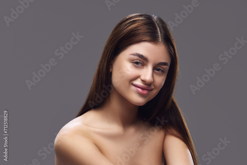 Young brunette woman with perfect, clean and smooth skin looking at camera against grey studio background. Concept of foundation makeup advertising to highlight matching skin tones, seamless coverage.