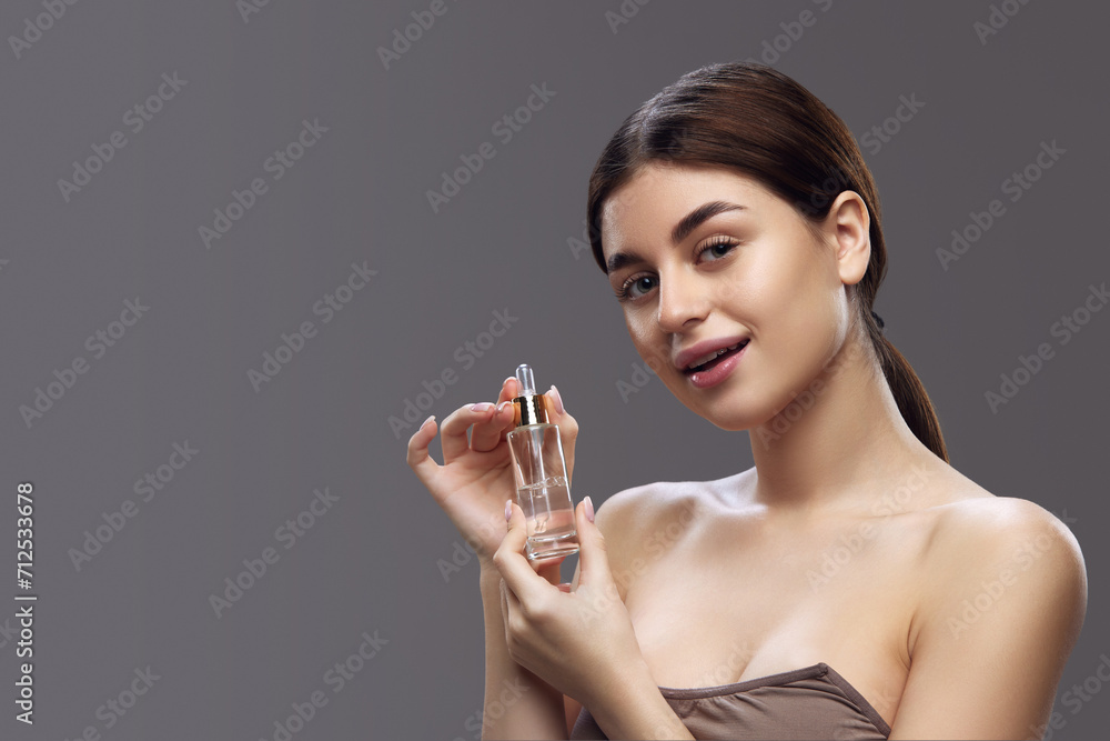 Young woman presenting a bottle of perfume, smiling subtly with focus on the product and her natural beauty. Concept of self care, organic and sustainable perfumes, cosmetic products. Young woman presenting a bottle of perfume, smiling subtly with focus on the product and her natural beauty. Concept of self care, organic and sustainable perfumes, cosmetic products.