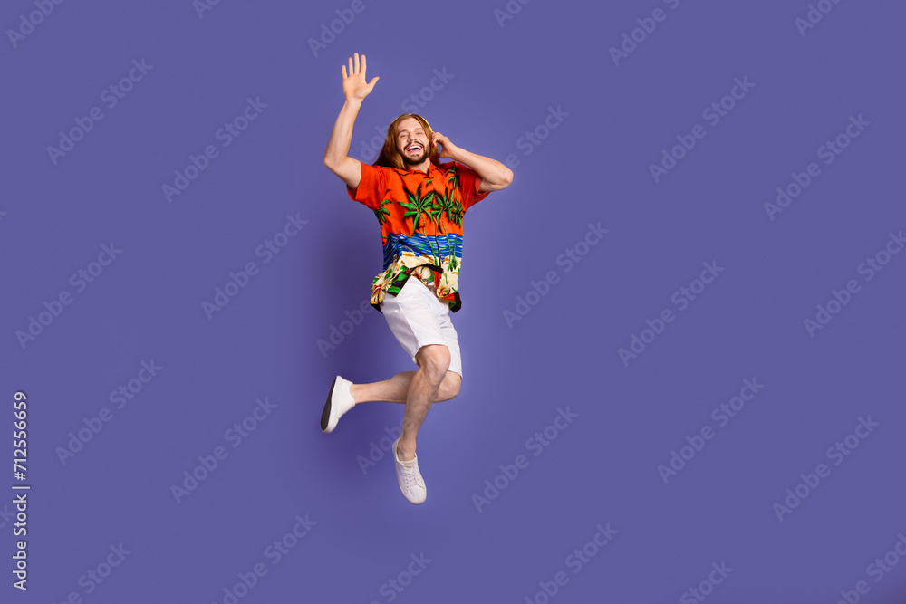 Full size photo of cheerful man wear print shirt white shorts touch headphones jumping waving palm isolated on purple color background Full size photo of cheerful man wear print shirt white shorts touch headphones jumping waving palm isolated on purple color background