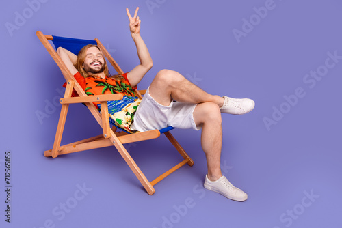 Full size photo of cheerful man wear print shirt sit on armchair showing v-sign on summer weekends isolated on purple color background