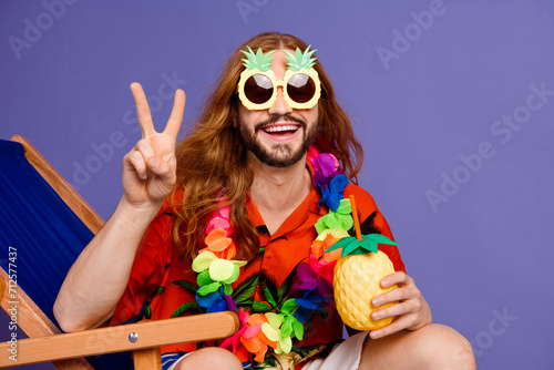 Photo of cheerful man with long hair in sunglass sit on armchair hold pineapple cocktail show v-sign isolated on violet color background