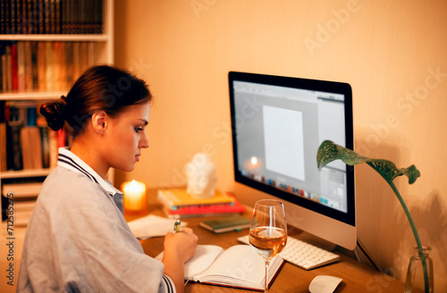 Pensive young female freelancer working on desktop computer from home while writing notes on agenda