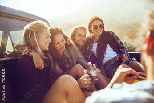Group of friends laughing and drinking beer at the back of a car on a road trip