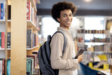Happy black male student with backpack on uses tablet in library, smiling at camera, showing the joy of mixing tech and traditional study methods