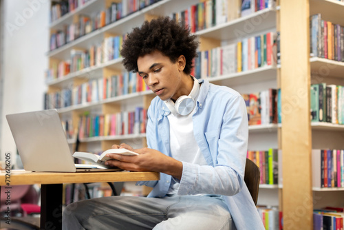 Focused black student guy sitting in library in front of laptop, diligently reading book, creating scene of concentrated study and blended learning methods