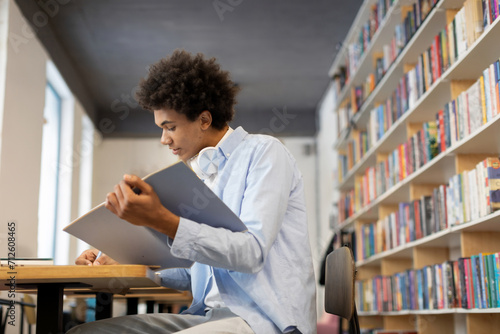 Black male student writing notes in copybook and holding book, encapsulating dedicated study session with blend of traditional and literary learning, sitting in library