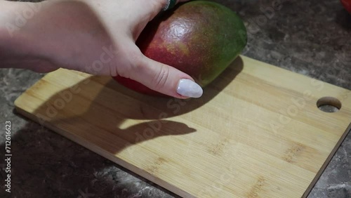 The girl takes a red mango with a pit and starts to cut it open. First she cuts off one side of the mango with a knife, then the other side of the mango. The pit of the mango remains in the middle. Th