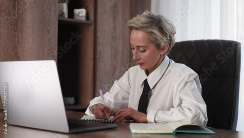 Stressed annoyed middle-aged business woman sitting at desk in office, writing on paper and crumpling it. Angry female entrepreneur not satisfied with work done. Unproductiveness, frustration, burnout photo