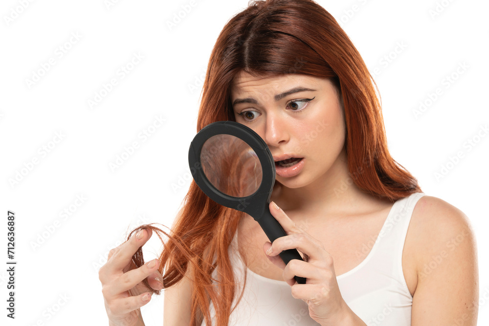 Worried redhead woman inspecting her hair with a magnifying glass on a white background Worried redhead woman inspecting her hair with a magnifying glass on a white background