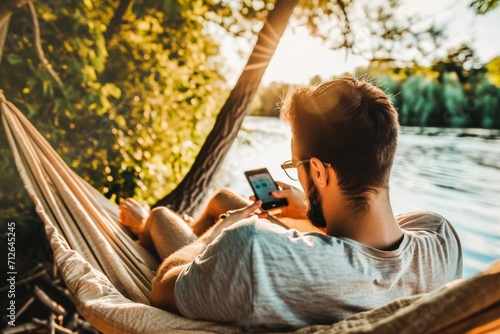 Man relaxing in a hammock by the lake, using a smartphone during a serene summer sunset.