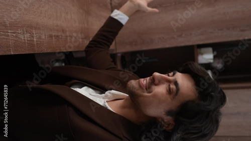 POV vertical video cheerful businessman in formal suit talking on video call, bragging about office using smartphone sitting in desk. Successful male entrepreneur communicates looking at camera.