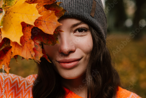 Caucasian woman walking outdoor, portrait of young european lady in warm sunny autumn park season, fall, hold yellow orange red leaves, dressed warm orange sweater