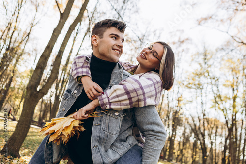 Heterosexual caucasian young loving couple, man carrying on back woman in sunny weather, hugging smiling kissing laughing spending time together. Autumn, fall season, orange yellow red maple leaves  © Volodymyr