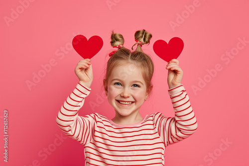 Happy child holding up red heart cutouts, symbolizing joy and love during Valentine's Day, isolated on pink