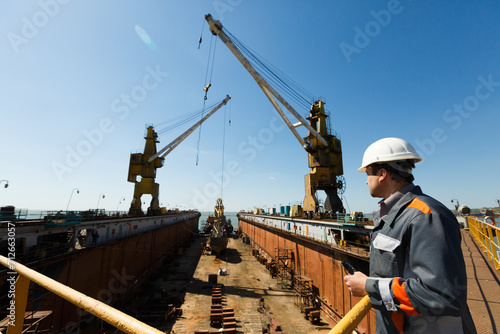 Professional overseeing extensive shipbuilding process, massive cranes erect vessel in dry dock against a clear sky. Maritime engineer in hardhat and reflective vest monitors construction.