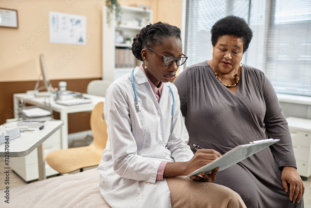 Obraz premium Medium full shot of Black woman physician in glasses holding clipboard and filling in medical record of senior female patient sitting in modern clinic office