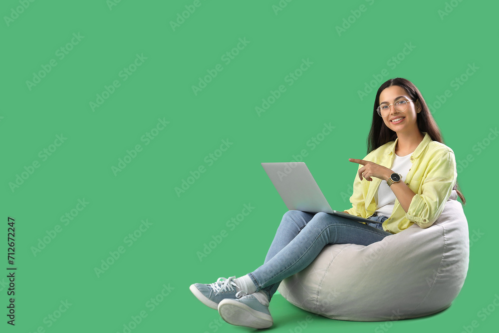 Young female programmer pointing at laptop while sitting on grey beanbag chair against green background Young female programmer pointing at laptop while sitting on grey beanbag chair against green background