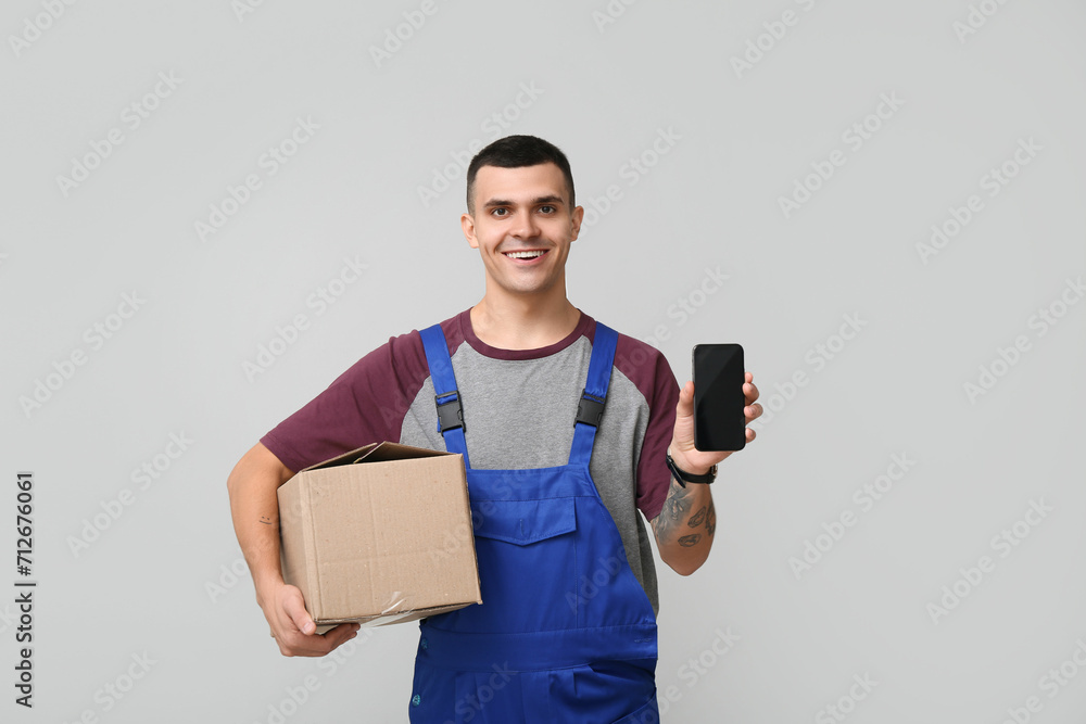 Young delivery man in blue uniform holding parcel and mobile phone with blank screen on grey background Young delivery man in blue uniform holding parcel and mobile phone with blank screen on grey background