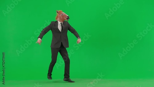 A man in a business suit with a horse head mask on a green studio background. A businessman appears in the frame in a comic manner, gallops like a horse and disappears. photo