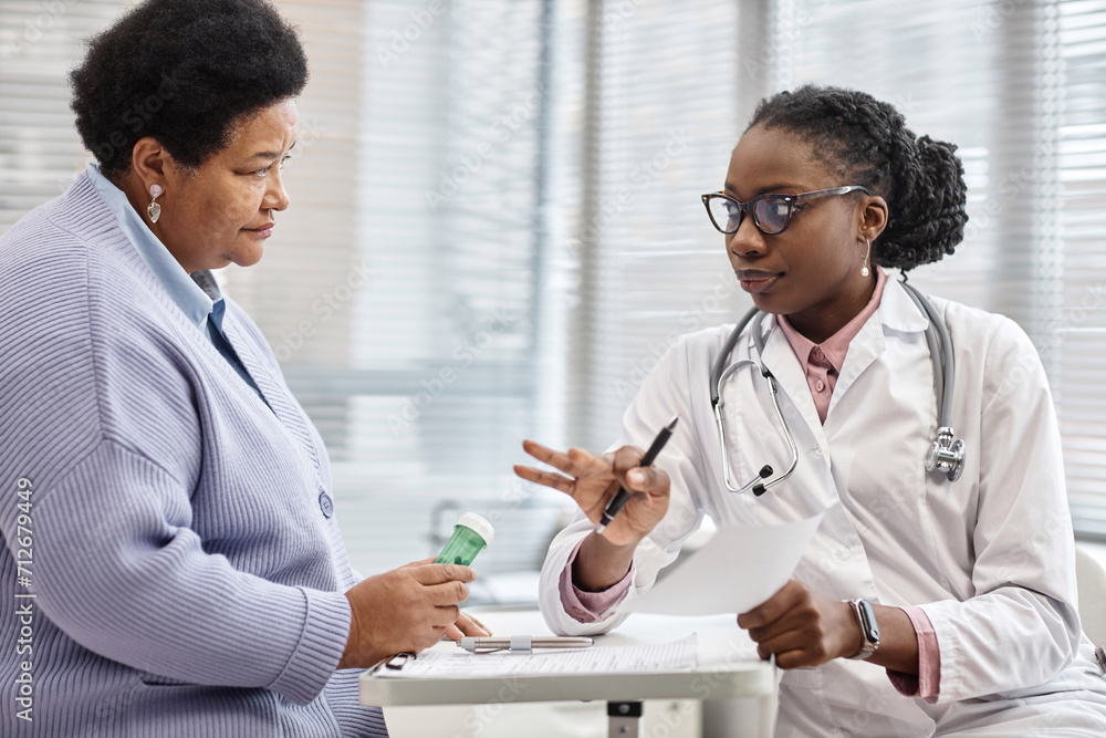 Fototapeta premium Senior African American female patient holding drug bottle while listening to doctors instructions about medication administration in clinic office