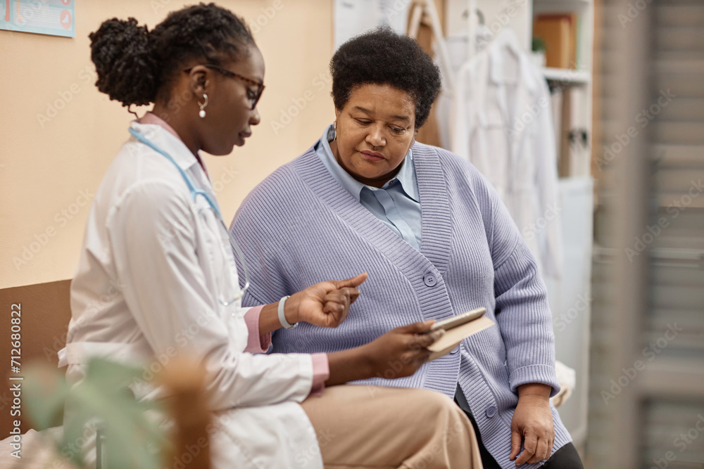 Obraz premium Senior African American woman looking at tablet in hand of female doctor talking about test results in clinic office