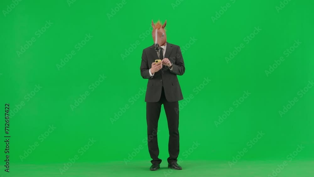 A man in a business suit with a horse head mask on a green studio background. A businessman is looking at something on his smartphone and is happy about good news or winning. Hard office work concept. A man in a business suit with a horse head mask on a green studio background. A businessman is looking at something on his smartphone and is happy about good news or winning. Hard office work concept.