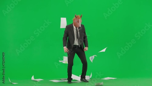 A man in a business suit with a horse head mask on a green studio background. The businessman looks through the documants, scatters them and begins to dance merrily. Hard office work concept.