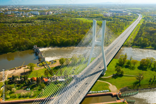 Cars drive on cable-stayed Redzinski Bridge over river flowing near scenic Wroclaw. Pylon bridge surrounded by lush green forests aerial motion along bridge