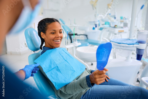 Happy black woman looking her teeth in mirror during appointment at dentist s office.