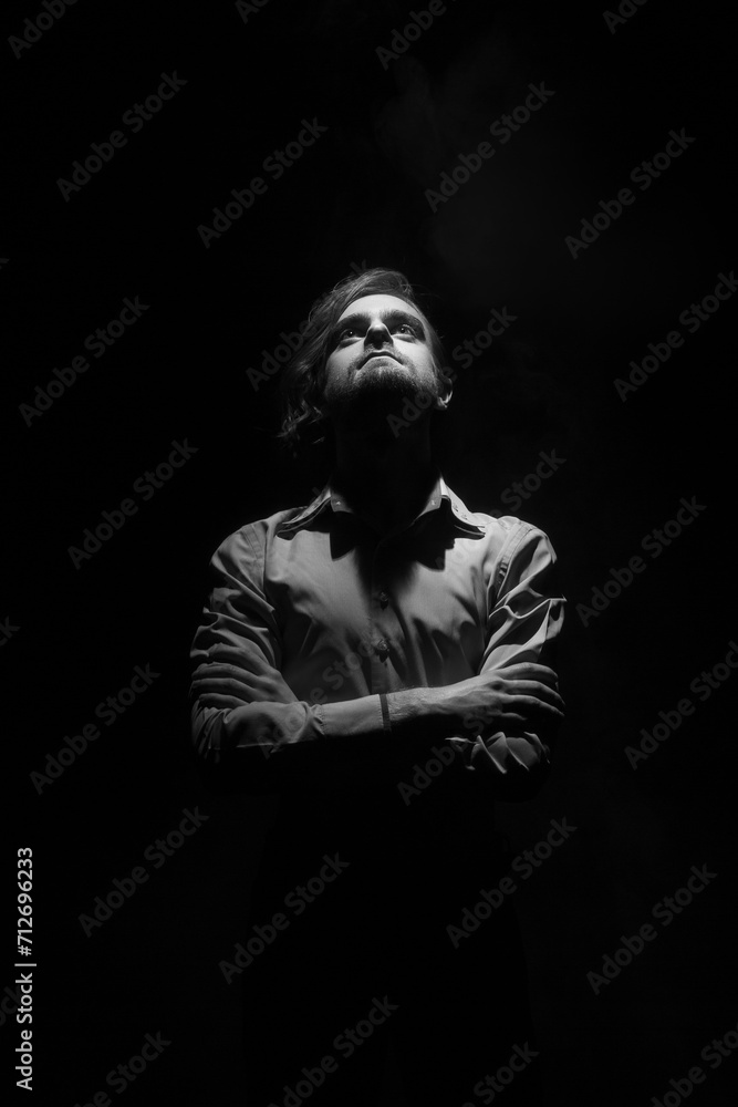 Black and white portrait of a young man with a beard, standing on a black background in a ray of light, crossing his arms over his chest and looking up