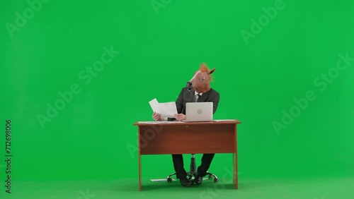 Man in business suit with horse head mask on studio green background. Businessman sitting at desk looking through documants and working at laptop. Heavy office work concept. photo