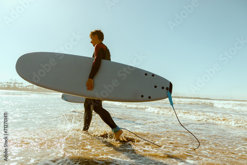 Back view of male surfer in wetsuit with his surfboard entering the sea. Surfing and ocean