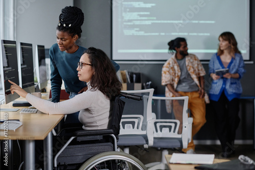 Black woman helping her colleague in wheelchair with code writing, coworkers standing on background photo