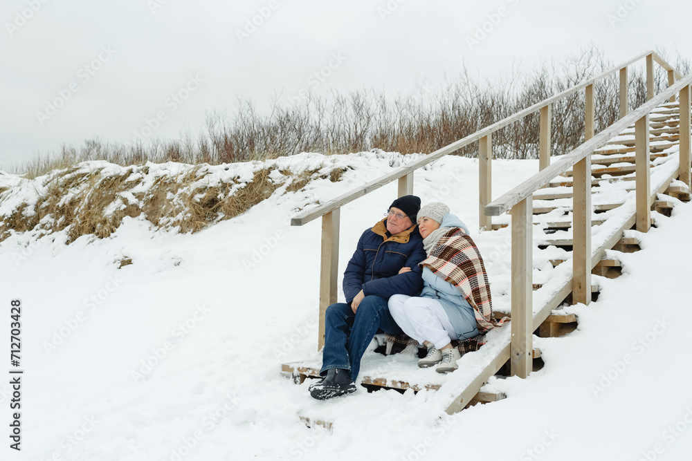 An elderly couple sitting together on a wooden staircase by the sea in winter.