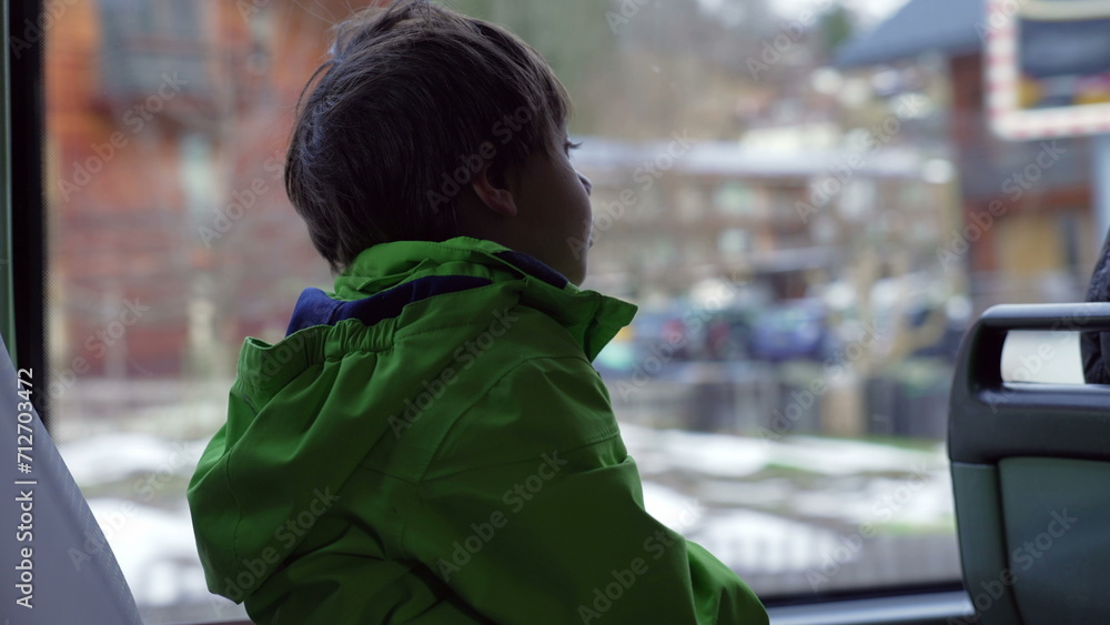 Obraz premium One small boy seated by bus window wearing green raincoat looking at view pass by inside transportation in motion