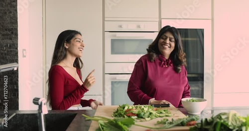 Indian 55s mom and 20s daughter hold hands, spinning, moving to favourite music dance together in modern cozy domestic kitchen spend carefree weekend at home, cooking, prepare healthy vegetarian dish photo