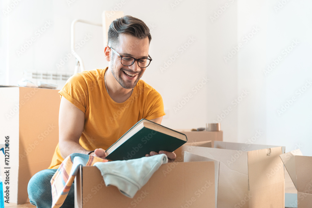 Young man unpacking cardboard boxes in new apartment. Excited man looking at the book packed inside a box. Young man unpacking cardboard boxes in new apartment. Excited man looking at the book packed inside a box.