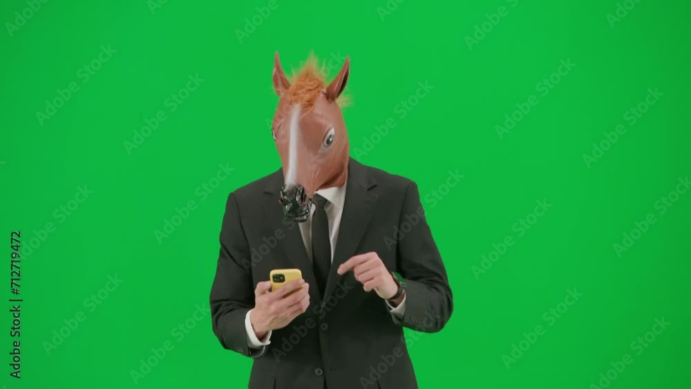 A man in a business suit with a horse head mask on a green studio background. A businessman is looking at something on his smartphone and is happy about good news or winning. Hard office work concept. A man in a business suit with a horse head mask on a green studio background. A businessman is looking at something on his smartphone and is happy about good news or winning. Hard office work concept.