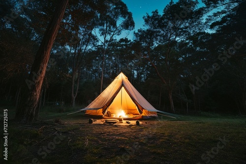Night and warm light exposure of an illuminated bell tent surrounded by trees and a small camp fire at night, outdoor camping lifestyle photo