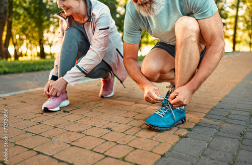Fototapeta Naklejka Na Ścianę i Meble -  Active retirement concept. Close up of caucasian senior man and woman in active clothes tying laces on sport sneakers at city park. Healthy married couple preparing for morning run during summer day.