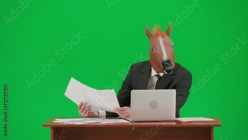 Man in business suit with horse head mask on studio green background. Businessman sitting at desk looking through documants and working at laptop. Heavy office work concept. photo