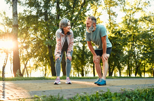 Recreation after workout. Happy old couple leaning with hands on knees and looking at each other while resting after outdoors jogging. Married man and woman taking care of health by common training. photo