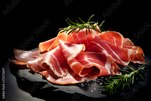 Prosciutto with fresh Rosemary served on Dark Stone cutting board. Prosciutto ham Slices with a sprig of fresh rosemary and salt. Italian food. Delicious Cured meat on dark backdrop. Italian food