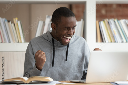 Overjoyed biracial guy sit at desk feel euphoric with good online news on laptop, college approval letter, excited african American male student triumph get positive decision email on computer