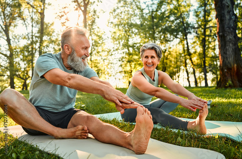 Flexible exercises for body. Sporty man and woman with grey hair stretching on yoga mats with hands to one leg during outdoors workout. Happy married couple with bare feet warming up together at park. photo