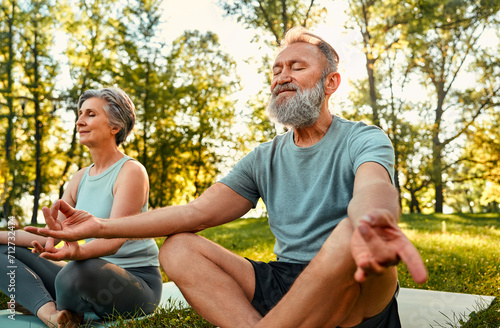 Yoga practice at park. Peaceful grey haired man and woman sitting in lotus pose with closed eyes and meditating on fresh air. Couple on retirement keeping hands in mudra gesture and feeling calmness. © HBS