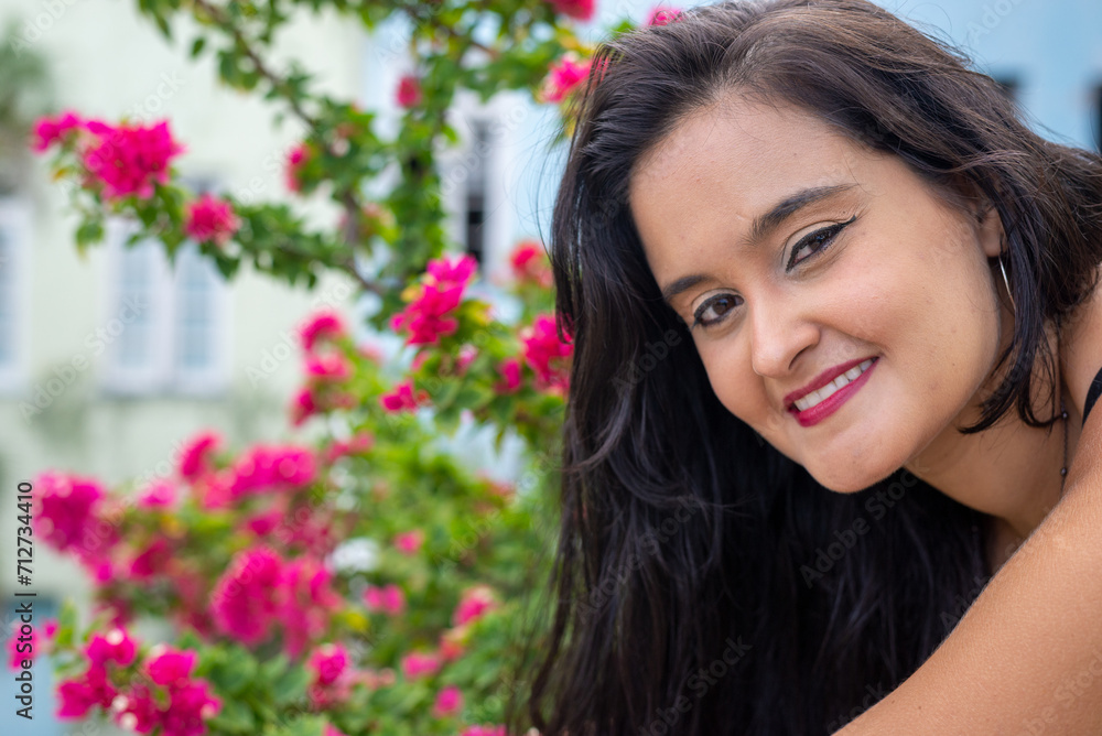 Portrait of a beautiful young woman, with long black hair, looking at the camera with flowers in the background.