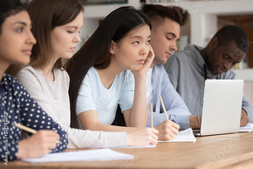 Attentive multiethnic young people sit in row at desk listen to teacher or tutor talking at classroom lecture, concentrated multiracial international students make notes at lesson, education concept